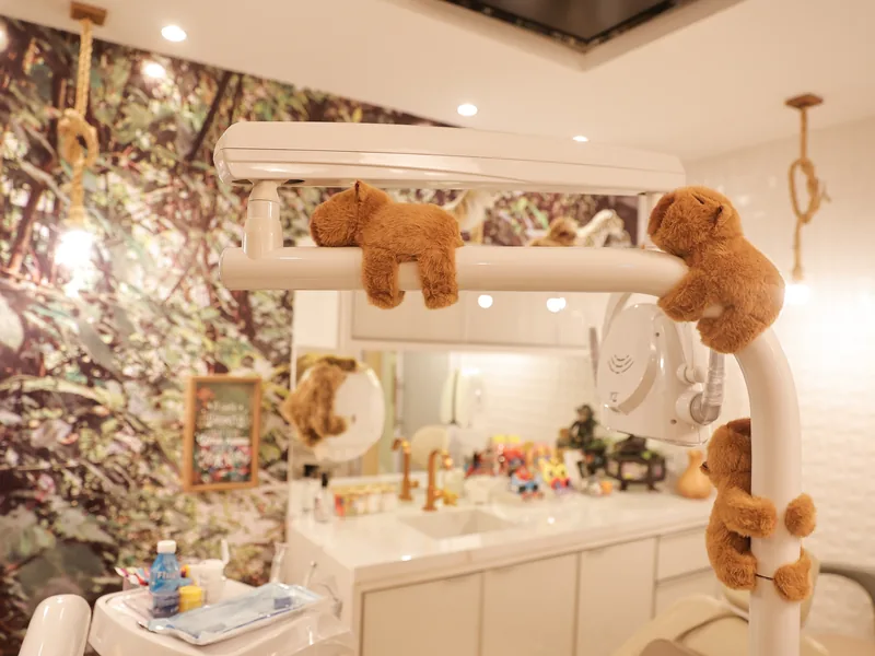 Consultório da Adelar Kids: refletor odontológico com macacos de pelúcia pendurados, parede com painel de floresta e bancada branca