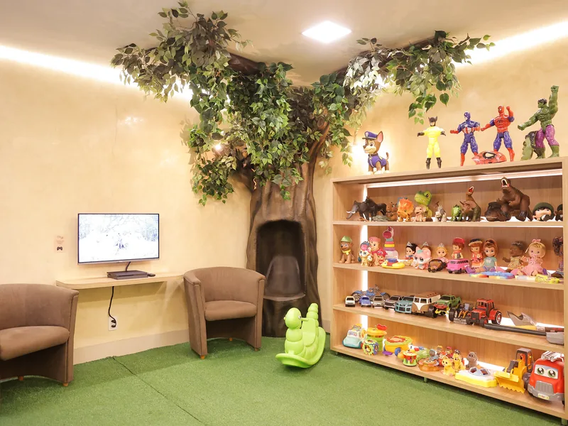 Espaço de espera da Adelar Kids com árvore cenográfica, poltronas para os acompanhantes, televisão e estante de brinquedos
